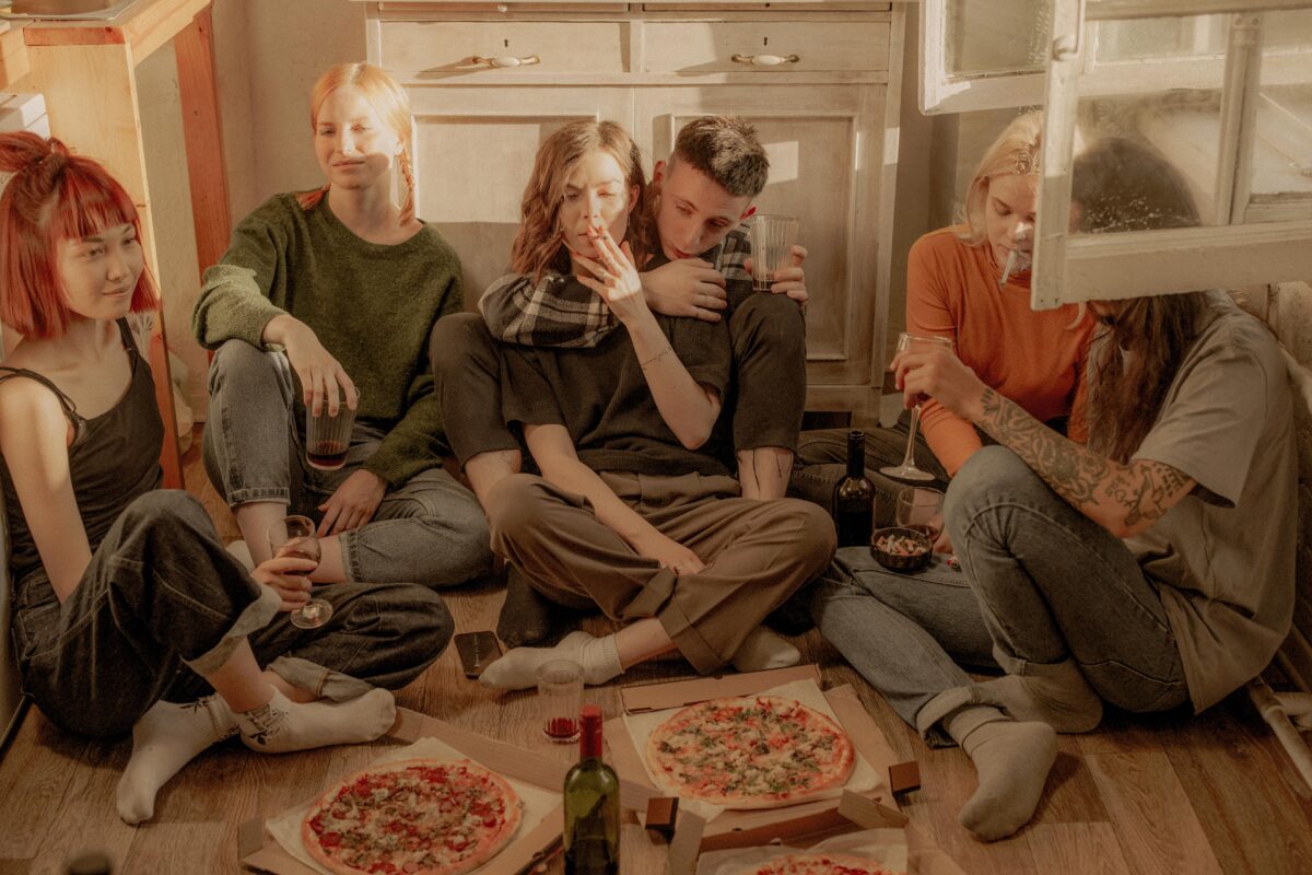 Groupe de personnes assises par terre. Certaines boivent un verre, d'autres fument. Il y a de la pizza devant elles.