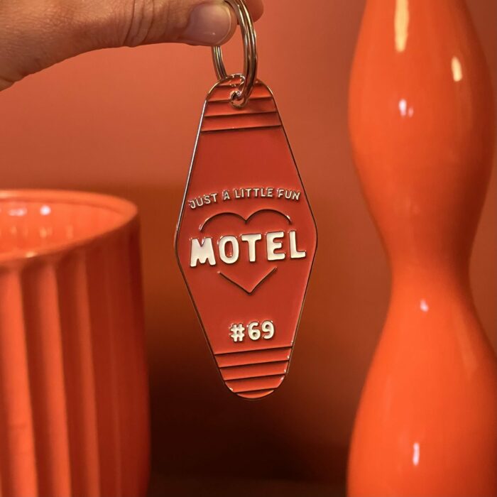Image d'une main tenant le porte-clé "Motel" de Just A Little Fun sur un fond rouge